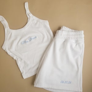May include: A white sleep set, comprising a vest top and shorts. The vest top features thin straps and the embroidered text "Mrs. Brown" in light blue. The shorts have an elasticated waistband and the embroidered text "06.10.26" in light blue.