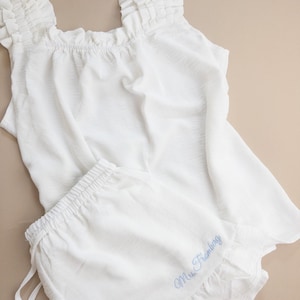 May include: White pajama set with a ruffled top and matching shorts. The top has ruffled straps and a square neckline. The shorts have a drawstring waist and a ruffled hem. The name "Mrs. Framberg" is embroidered on the shorts in light blue.