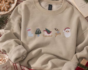 Könnte beinhalten: Ein beigefarbenes Sweatshirt mit Rundhalsausschnitt und kleinen, mittig platzierten Weihnachtsmotiven als Stickerei, darunter ein Strumpf, ein Tannenbaum, ein Schlitten, ein Rentier und ein Weihnachtsmann-Gesicht. Es hat lange Ärmel, einen Rippkragen und ein Gildan-Etikett.