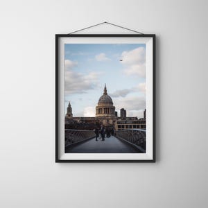 Puede incluir: Impresión enmarcada de la Catedral de San Pablo en Londres, Inglaterra. La imagen captura la icónica cúpula y arquitectura desde un puente, con gente caminando. El cielo es azul con nubes dispersas, y el tono general es apagado.