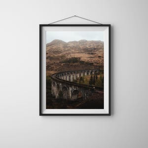 Può includere: Stampa incorniciata con un paesaggio che presenta un ponte in pietra curvo. Il ponte attraversa una valle con una catena montuosa sullo sfondo. La tavolozza dei colori è attenuata con toni terrosi.