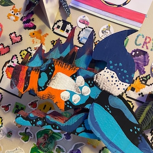 May include: A large, colorful, handmade papercraft dragon claw with blue, orange, and black accents. The claw is decorated with white polka dots and is surrounded by pixel art stickers and drawings.