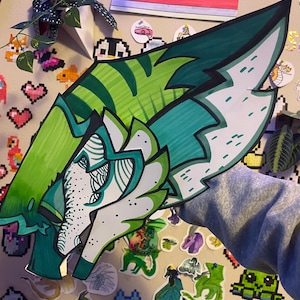 May include: A large, hand-drawn, green and white dragon head with intricate details. The artwork features shades of green and white, with a stylized design. The dragon head is the focal point, surrounded by various stickers and decorations.