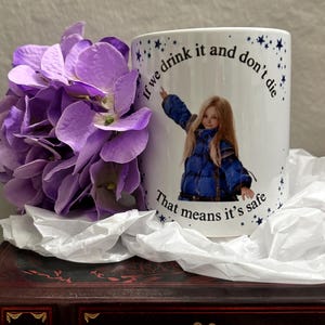 May include: A white ceramic mug featuring a graphic of a person in a blue puffer jacket with the text, "If we drink it and don't die, That means it's safe," surrounded by small dark blue stars, positioned next to a cluster of purple hydrangea flowers.