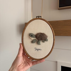 May include: An oval wooden embroidery hoop containing a light beige fabric background. A needle-felted turtle with a brown shell and sage green limbs is centered on the fabric. Below the turtle, the name Tason is embroidered in black script.