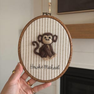 Può includere: Un telaio da ricamo ovale in legno con una scimmia in feltro marrone e beige su uno sfondo di tessuto a righe. Il nome "Hawke Michael" è ricamato sotto. Il diametro del telaio è di circa 15 cm.