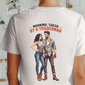 K&ouml;nnte beinhalten: Eisgraues T-Shirt mit einer Grafik eines Paares und dem Text "WARNING: TAKEN BY A TRADESMAN" in Orange und Schwarz. Die Illustration zeigt einen Mann und eine Frau in Freizeitkleidung.