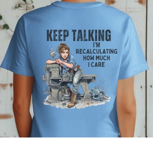 Puede incluir: Camiseta azul claro con el texto "KEEP TALKING I'M RECALCULATING HOW MUCH I CARE." El gráfico presenta a una mujer apoyada en una máquina. Lleva una camisa vaquera, vaqueros y botas de trabajo. La camiseta es de cuello redondo.