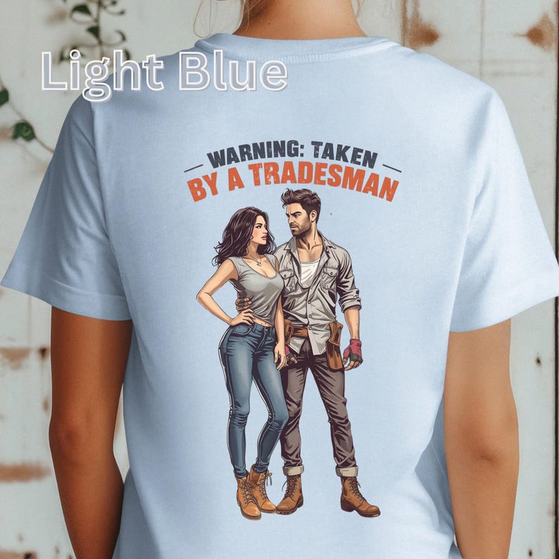 K&ouml;nnte beinhalten: Hellblaues T-Shirt mit dem Text "WARNING: TAKEN BY A TRADESMAN" &uuml;ber einer Illustration eines Paares. Die Grafik zeigt einen Mann in Arbeitskleidung und eine Frau in Freizeitkleidung.