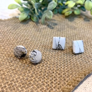 May include: A collection of white and black stone earrings. Two round stud earrings and two square stud earrings are displayed on a burlap surface. The earrings have a marbled pattern. Green foliage is in the background.