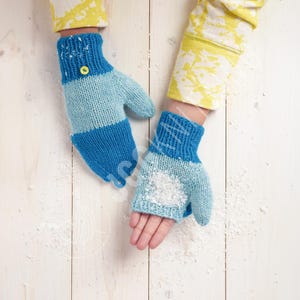 Könnte beinhalten: Gestrickte Fäustlinge in Blautönen, mit einem hellblauen Streifen und einer dunkelblauen Manschette. Ein Fäustling hat einen gelben Knopf. Die Fäustlinge liegen auf einer weißen Holzoberfläche mit weißen Flocken.