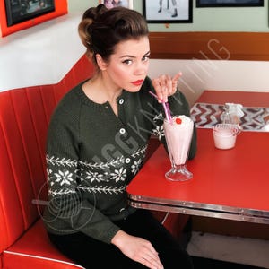 Może przedstawiać: Kobieta w ciemnozielonym kardiganie z białymi wzorami płatków śniegu siedzi przy czerwonym stole w barze. Trzyma słomkę w szklance koktajlu mlecznego z bitą śmietaną i wiśnią. Kardigan ma białe guziki.