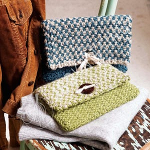 May include: Hand-knitted clutch purses in blue, green, and grey. The top purse is blue and white, the middle one is green and white, and the bottom one is grey. Each purse has a unique texture and design, with a button closure. The purses are stacked.