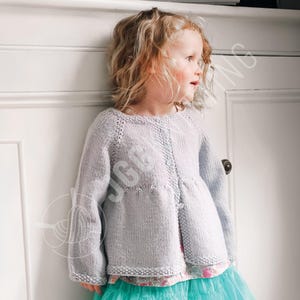 Könnte beinhalten: Hellgraue Strickjacke mit langen Ärmeln und rundem Ausschnitt. Die Strickjacke hat eine Knopfleiste vorne und einen leicht ausgestellten Saum. Das Kind trägt einen türkisfarbenen Rock und ein Unterhemd mit Blumenmuster.