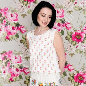 Op de afbeelding: Een mouwloze witte gebreide top met een bloemenpatroon van roze bloemen en groene stelen. De top heeft een ronde halslijn en een getailleerde taille. De achtergrond is een bloemenbehang met grote roze rozen.