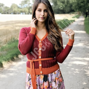 Könnte beinhalten: Ein roter Cardigan mit orangefarbenen Verzierungen und einem passenden Gürtel. Der Cardigan hat einen V-Ausschnitt und ist zugeknöpft. Die Ärmel und der Kragen sind orangefarben eingefasst. Das Model trägt einen Rock mit Blumenmuster.