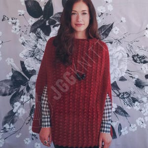 May include: A red knitted poncho with a textured pattern and a single black button closure. The poncho is worn over a checkered shirt. The image has the text "JGGKNIT" in the lower right corner.