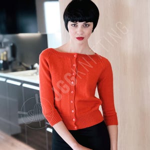 Puede incluir: Un cárdigan naranja con mangas tres cuartos y cierre de botones. El suéter tiene un dobladillo y puños acanalados. La modelo lleva una falda negra. El fondo incluye una cocina y una pared de madera.