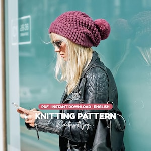 Puede incluir: Un gorro de punto color magenta intenso con un patrón de puntos texturizados y un gran pompón a juego en la parte superior. El gorro cuenta con un borde acanalado y se presenta como un patrón de tejido PDF en inglés, Create with joy.