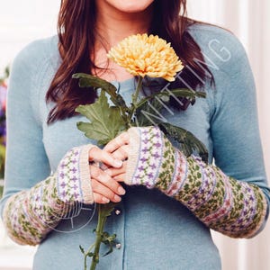 Könnte beinhalten: Gestrickte Armstulpen mit Fair-Isle-Muster in Grün, Lila und Rosa. Die Armstulpen werden von einer Person gehalten, die einen hellblauen Pullover trägt und eine gelbe Blume hält.