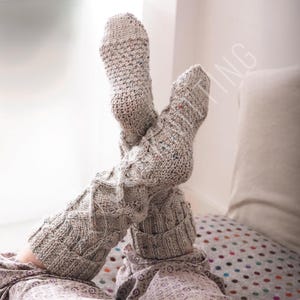 May include: Cozy, hand-knitted gray leg warmers with a textured diamond pattern. The warmers are speckled with flecks of color and are shown with the feet up, suggesting comfort and relaxation. Perfect for cold weather or lounging.