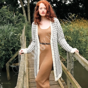 Op de afbeelding: Een lange, open cardigan met een pied-de-poule patroon in grijs en wit, gedragen over een bruine jurk. De cardigan heeft witte randen en wordt gecombineerd met een dunne zwarte riem. De scène speelt zich buiten af op een houten brug.