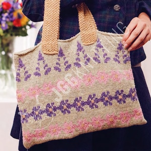 Puede incluir: Bolso tote tejido beige con estampados florales rosas y morados. El bolso tiene asas color canela y forma rectangular. El diseño floral incluye flores estilizadas y patrones de hojas. El bolso lo sostiene una persona que lleva una blazer azul oscuro.