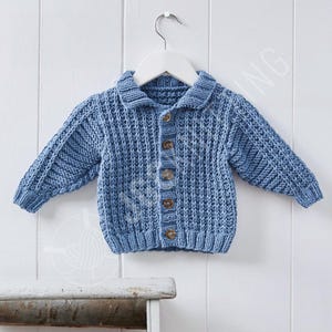 May include: A light blue, knitted baby cardigan with a collar and long sleeves. The cardigan has a ribbed texture and is fastened with five brown buttons. It hangs on a white hanger against a white wooden background.