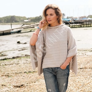 Peut inclure: Un poncho en tricot beige clair avec un large col et des manches courtes. Le poncho présente un motif rayé horizontal texturé. La personne sur l'image porte un jean bleu. L'arrière-plan est une plage avec des bateaux.
