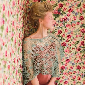 May include: A sage green crochet poncho featuring an open net pattern with small leaf-shaped motifs throughout. The garment has a wide, loose fit with a boat neckline and short, flowing sleeves, worn over a solid-colored, sleeveless undergarment.