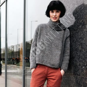 Puede incluir: Un suéter gris de gran tamaño con un cuello alto y doblado. El suéter se combina con pantalones de color óxido. La modelo está apoyada contra una pared de piedra texturizada junto a una ventana de cristal.