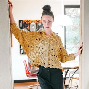 May include: Mustard yellow knitted cardigan with a hood and button closure. The cardigan features a textured knit pattern with openwork details. The sleeves are wide and the garment is worn over black pants.