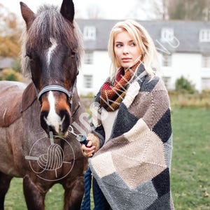May include: A woman wrapped in a patchwork blanket stands beside a brown horse. The blanket has a geometric pattern in grey, brown, and black. The woman wears a patterned scarf and a light-coloured sweater. The horse has a white blaze.