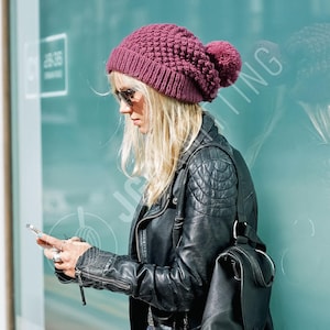 May include: A knitted, claret beanie hat with a large pom-pom. The hat is worn with a black leather jacket and sunglasses. The person is holding a smartphone.