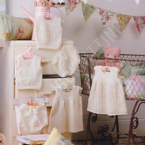 May include: A collection of cream-colored knitted baby garments including sleeveless vests, a dress, and bloomers. The items feature delicate openwork patterns and small embroidered details, displayed on hangers and a small chair with a pink teddy bear.