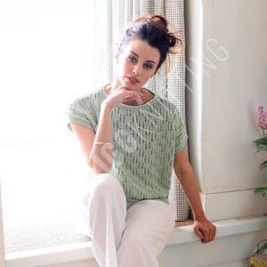以下が含まれることがあります： テクスチャードデザインの、ライトグリーンの半袖ニットトップス。白いパンツと組み合わせています。女性は窓枠に座っています。手首にはシルバーのブレスレットがあります。