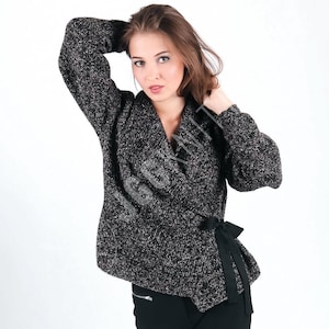 May include: A black and white speckled wrap sweater with a black tie belt. The sweater has long sleeves and a shawl collar. The text "JGGKNIT" is visible on the sweater. The model is wearing black pants.