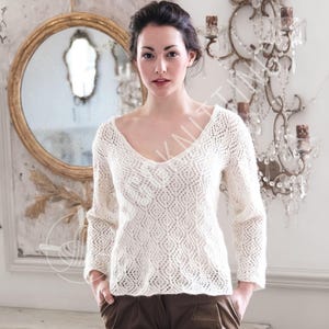 Può includere: Maglione color crema a maniche lunghe con scollo a V profondo e motivo a maglia a rombi. Il maglione è indossato con pantaloni marroni. Lo sfondo include uno specchio con cornice dorata e un lampadario di cristallo.