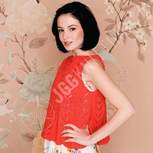 Op de afbeelding: Een mouwloze, koraalkleurige gebreide top met een geschulpte zoom. De top heeft een chevronpatroon en wordt gedragen over een kledingstuk met bloemenprint. De achtergrond is een bloemenbehang.