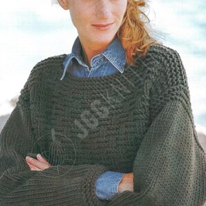 May include: A dark olive green knit sweater with a textured pattern. The sweater has a wide neckline and long sleeves. The person in the image is wearing a denim shirt underneath the sweater.