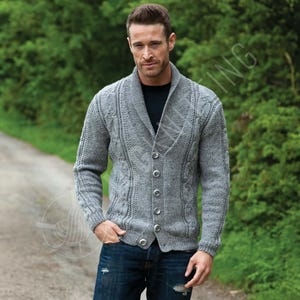 May include: A grey cable-knit cardigan with a shawl collar and button fastening. The knitted sweater has detailed patterns and is worn with blue jeans. The background is a blurred green outdoor scene.