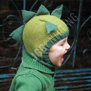 May include: A green and light green knitted dinosaur hat with matching neck warmer. The hat features triangular spikes along the top and a face opening. The neck warmer is a solid green color. The hat and neck warmer are made of a soft, knitted material.