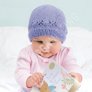 May include: A lavender knitted baby hat with a decorative openwork pattern near the brim. The baby is wearing a pink shirt and holding a book titled "Tom Kitten can feel..." The hat is a soft, cozy accessory for infants.