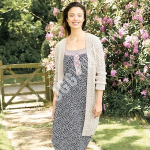 May include: A woman wearing a long, open-knit beige cardigan over a navy blue and white patterned dress with a pink and white bodice. The woman is smiling in a garden setting with a wooden gate and pink flowering bushes.