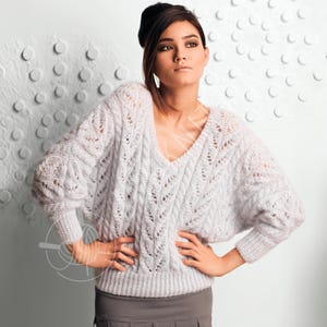 Puede incluir: Un suéter de punto abierto gris claro con cuello en V profundo y mangas largas. El suéter presenta un diseño texturizado y estampado y se usa con una falda gris. La modelo lleva un accesorio para el cabello negro.