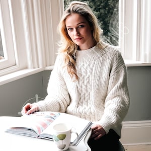 Puede incluir: Suéter de punto trenzado color crema con cuello redondo. El suéter tiene un patrón texturizado y mangas largas. Una mujer lleva el suéter mientras está sentada en una mesa con un libro y una taza.