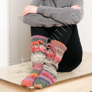May include: A pair of colorful, knitted leg warmers with a striped pattern in shades of pink, orange, purple, and green. Each leg warmer has a drawstring closure at the top. The leg warmers are resting on a wooden surface.