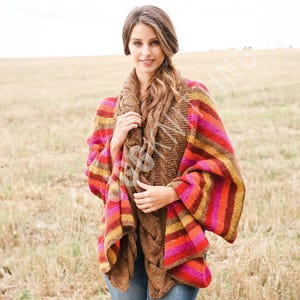 Puede incluir: Una mujer con un chal de punto marrón y un poncho a rayas de colores. El poncho presenta rayas horizontales en tonos rojos, naranjas y amarillos. El fondo es un campo de hierba seca.
