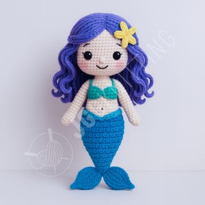 Pode incluir: Boneca de sereia de crochê feita à mão com cabelo azul e uma flor amarela. A boneca tem um top de biquíni turquesa e uma cauda azul. A boneca é feita de fio e tem um rosto sorridente.