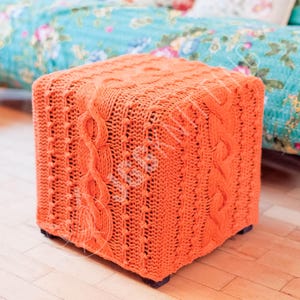 May include: An orange knitted cube-shaped ottoman with a cable knit design. The ottoman is made of a thick, textured yarn and sits on small black feet. The background features a floral patterned sofa.